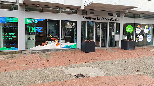 Außenansicht des Stadtwerke ServiceCenters mit großflächigen Schaufenstern, die farbenfrohe Werbegrafiken des Unternehmens TKRZ präsentieren. Links zeigt das Schaufenster das TKRZ-Logo mit dem Slogan 'TKRZ packt’s an!' und eine bunte Illustration von Tieren, die symbolisch für verschiedene Tarife stehen. Es sind Informationen zur 'Glasfaserstadt Emsdetten' und Standorte wie Emsdetten und Hauenhorst Museum abgebildet. Rechts vom Haupteingang befinden sich weitere grüne Grafiken mit Botschaften zum Engagement für Glasfaserausbau und regionalen Service. Der Boden vor dem Gebäude ist mit Pflastersteinen gestaltet und vermittelt ein einladendes Ambiente.