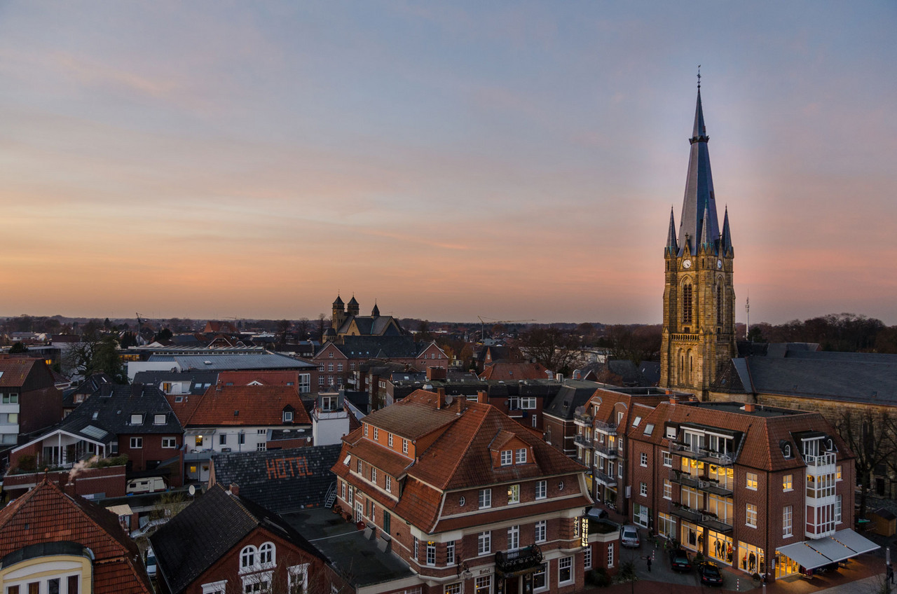 Stimmungsvolle Abendaufnahme einer deutschen Kleinstadt mit historischen Gebäuden und einer großen Kirche mit spitzem Turm, der die Skyline dominiert. Die Dächer der umliegenden Gebäude sind überwiegend mit roten Ziegeln gedeckt. Im Hintergrund erstreckt sich eine flache Landschaft unter einem sanften, pastellfarbenen Himmel während des Sonnenuntergangs. Die Szene vermittelt eine ruhige und idyllische Atmosphäre.